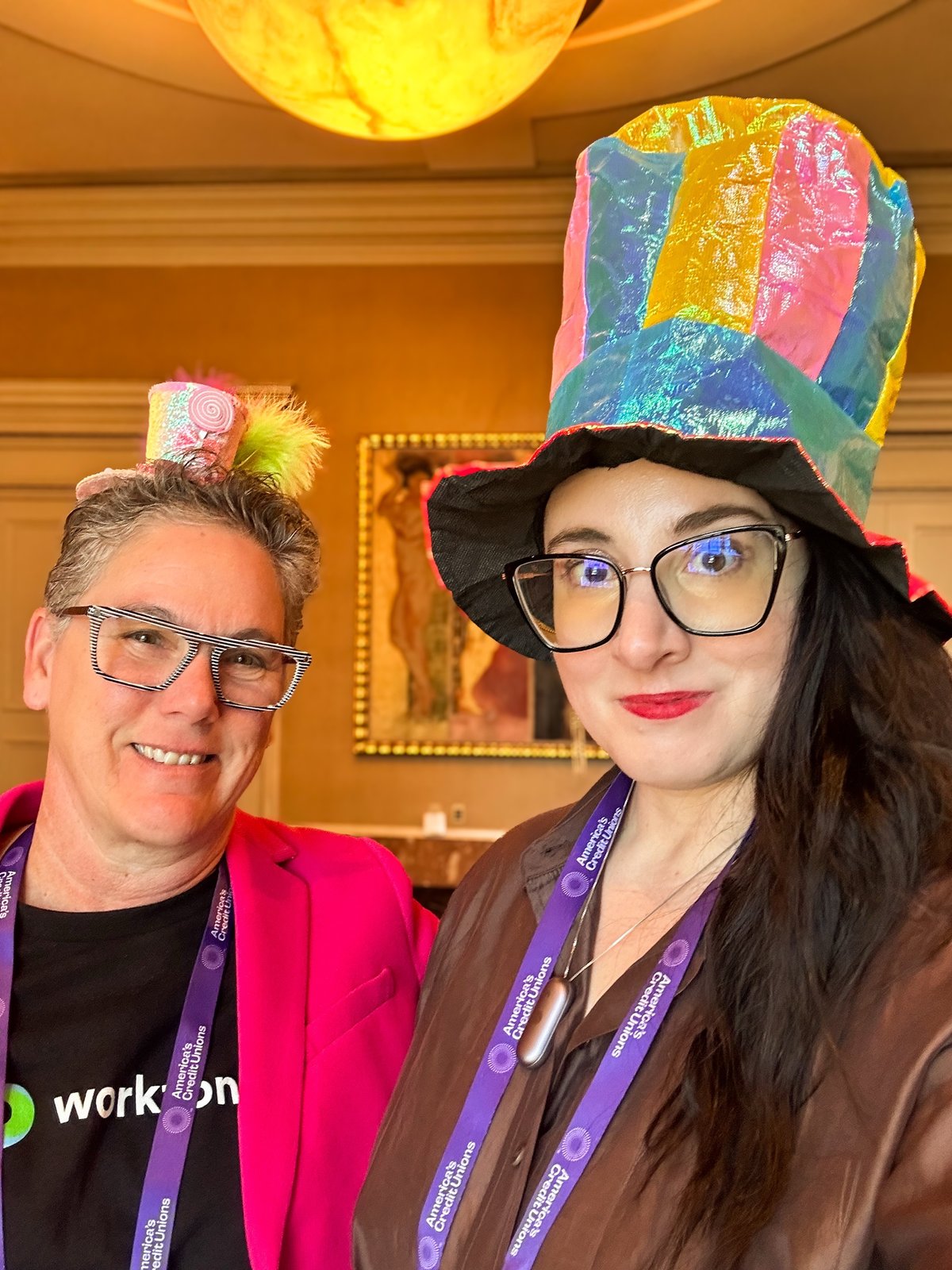 Two people in rainbow top hats and Workzone lanyards at a conference