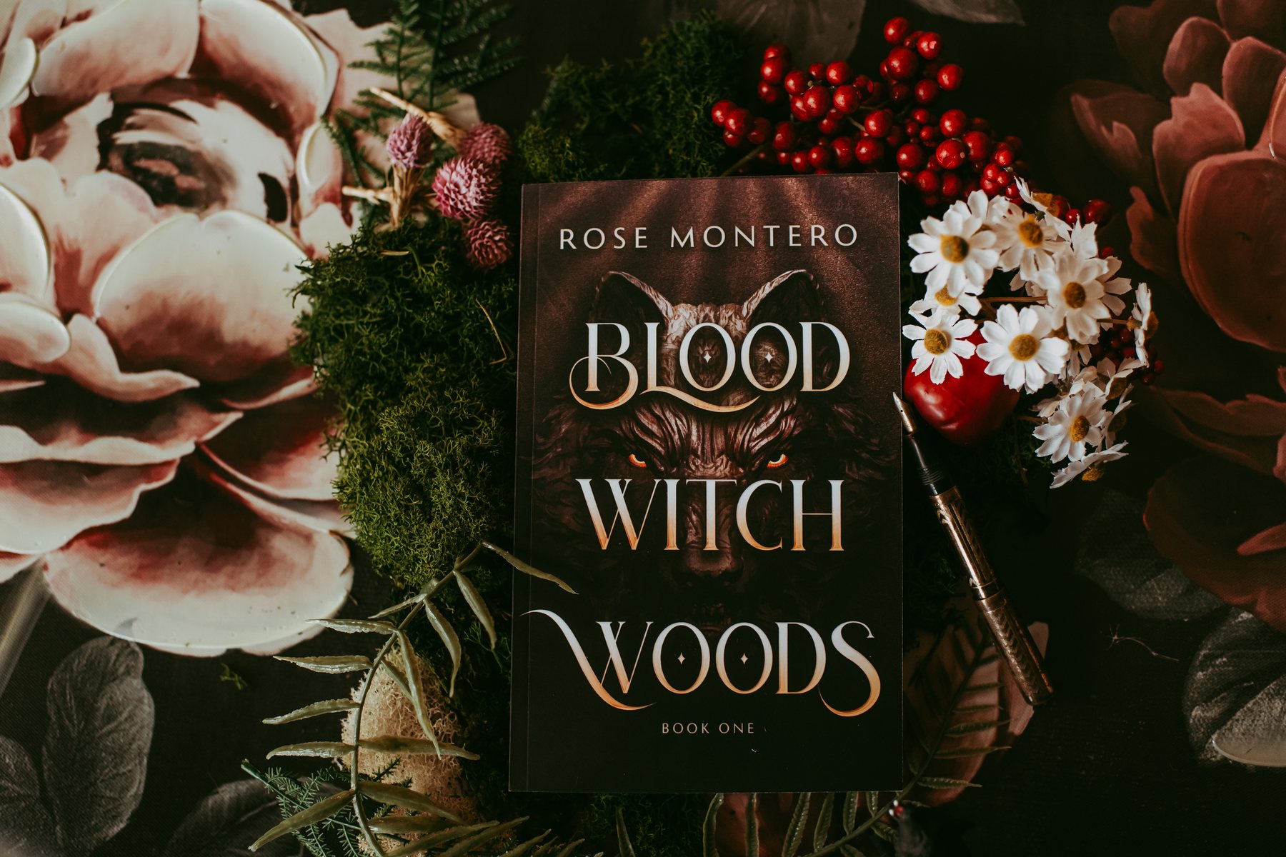 The cover of Blood Witch Woods by Rose Montero laid flat among moss, daisies, red berries and a fountain pen.