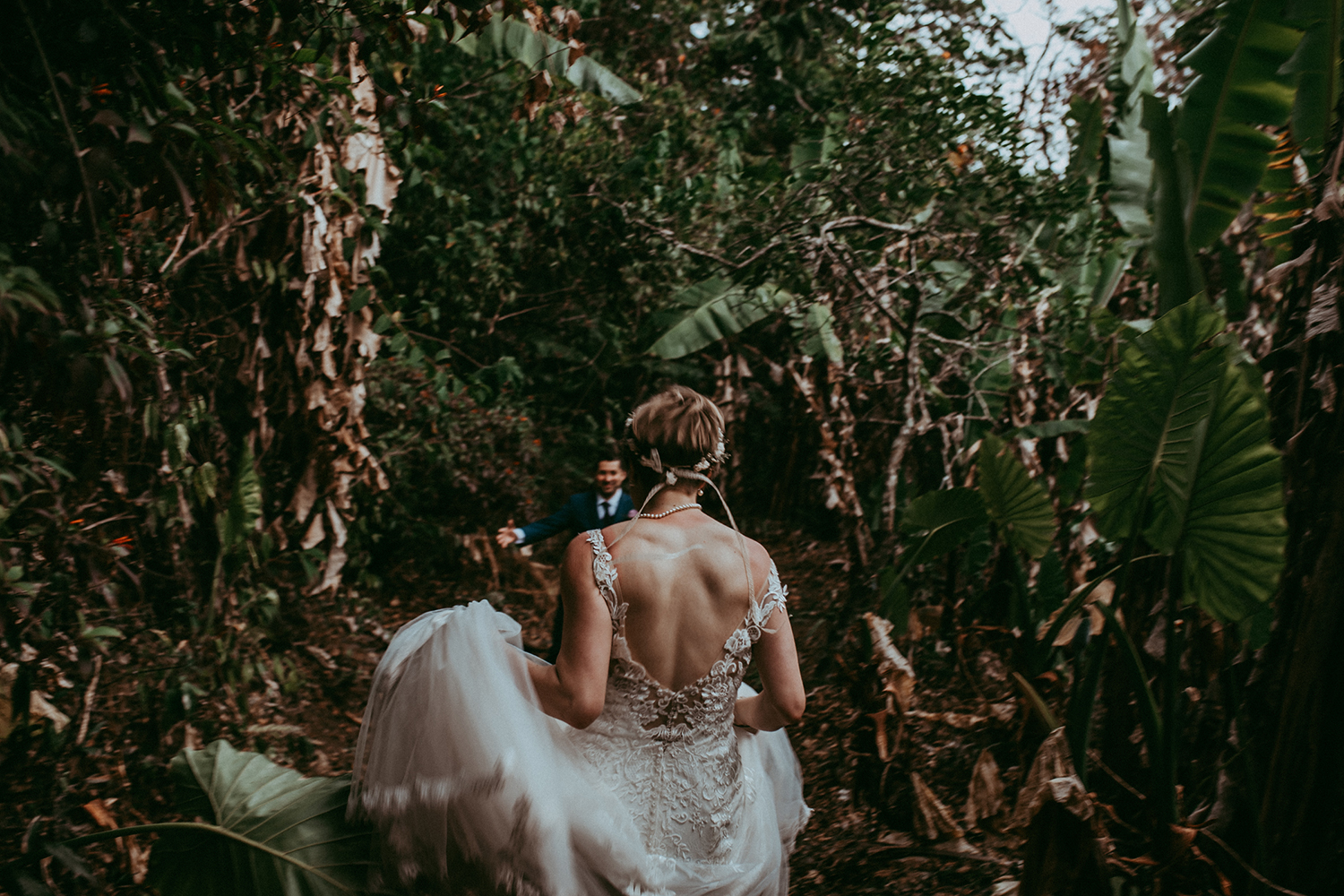 Bride in a lace backless dress walking through dense green foliage toward her groom