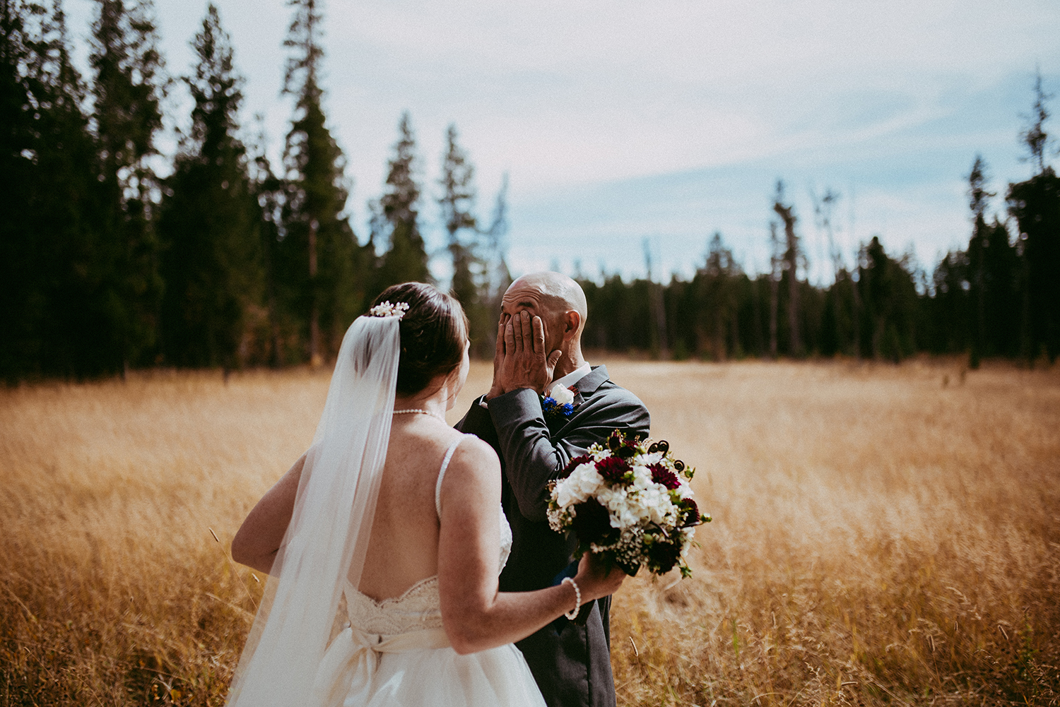 Bride in a veil holding a bouquet as the groom covers his face, overcome, in a golden meadow at a first look
