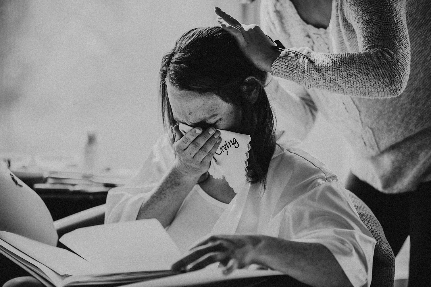 Black and white photo of a bride in a robe crying as she reads a letter while someone fixes her hair