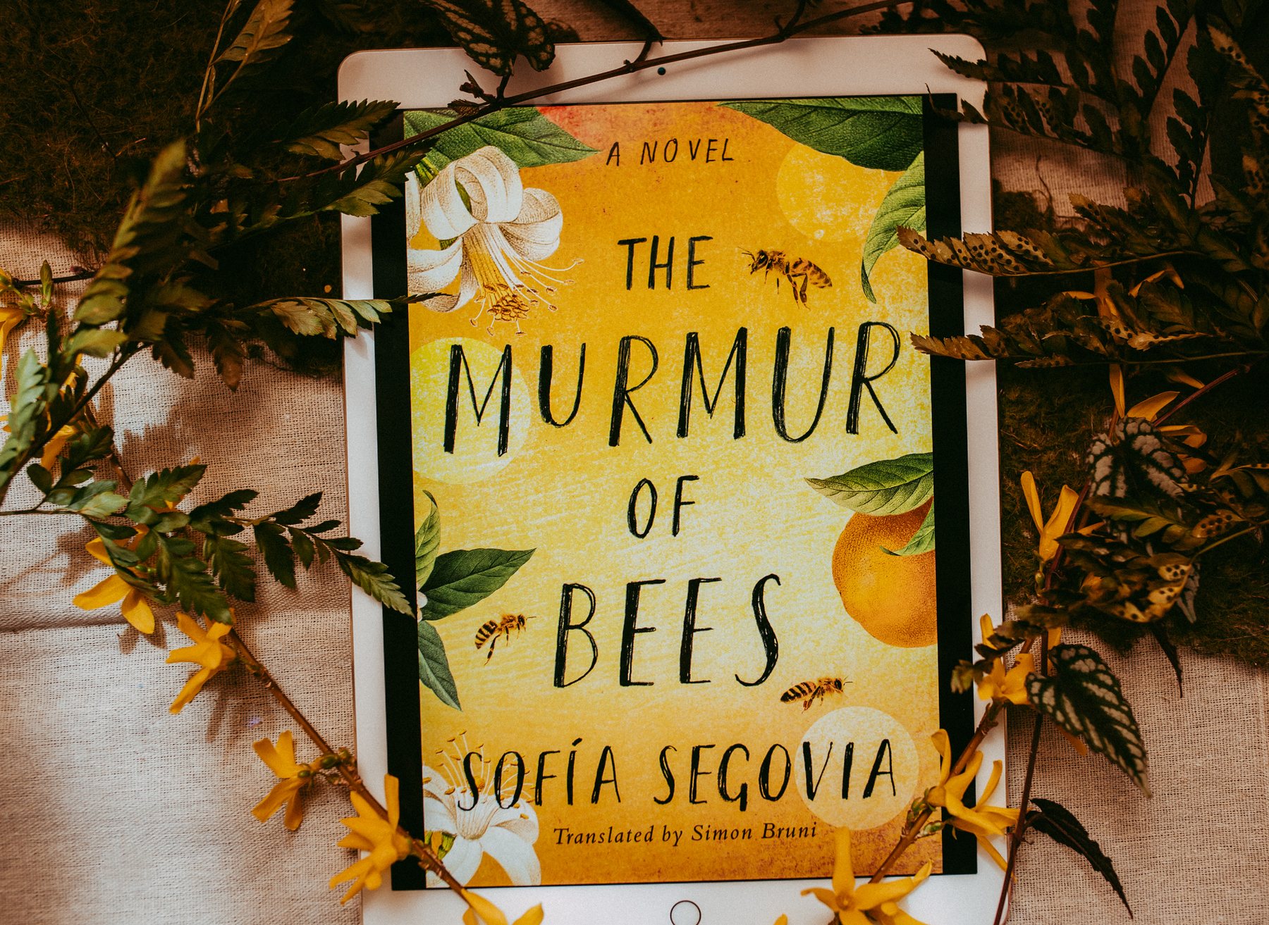 A tablet showing the cover of The Murmur of Bees by Sofia Segovia, laid on cloth with fern fronds and yellow flowers.