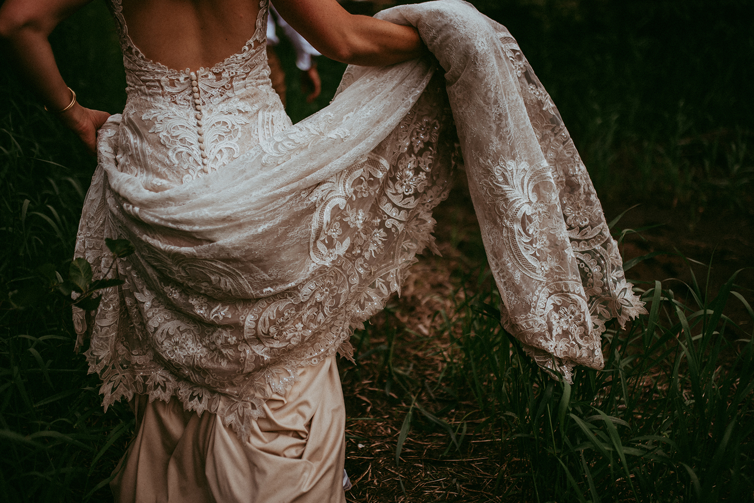 Close-up of a bride lifting the long lace train of her gown over dark grass