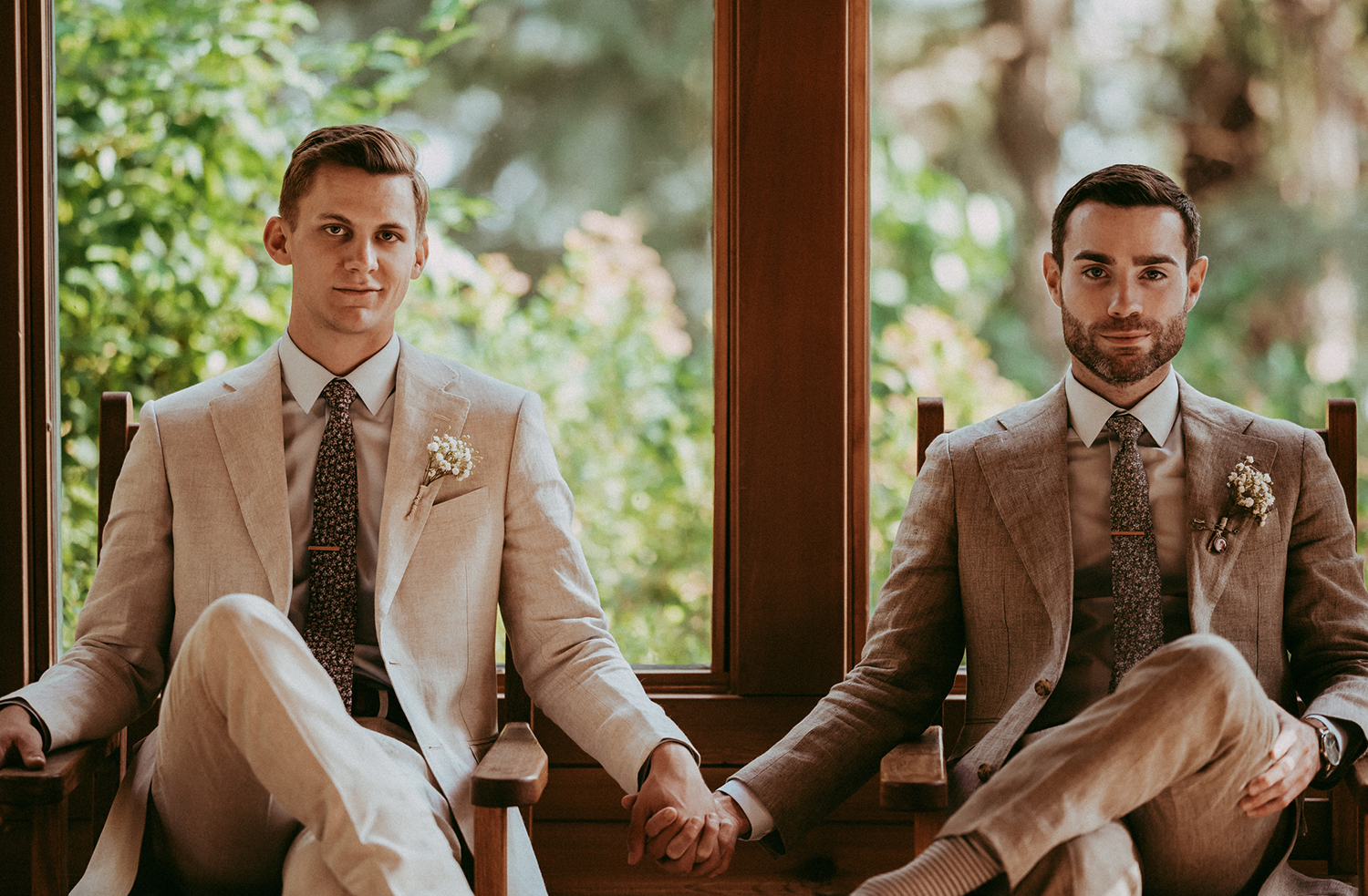 Two grooms in tan suits sitting side by side holding hands in front of a garden window